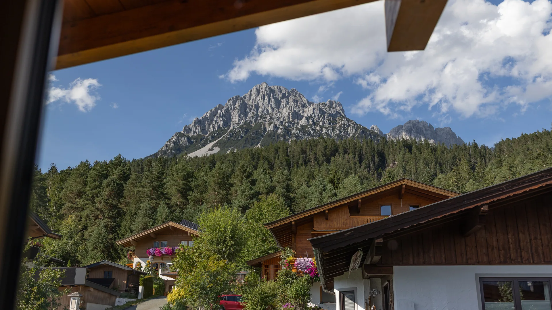 Blick aus dem FALKENSITZ Apartment auf den Wilden Kaiser — Holzhäuser im Vordergrund, Bergspitze unter blauem Himmel